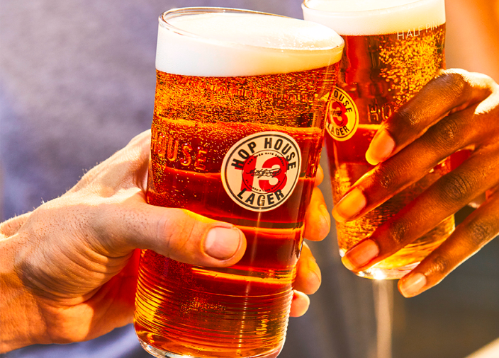 Zwei Hände, die ein Glas Hop House 13 Lager "anstoßen".