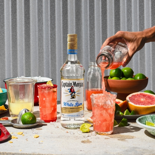 Bottle of Captain Morgan White Rum and two iced glasses of cocktail on a table with a hand filling one of the glasses