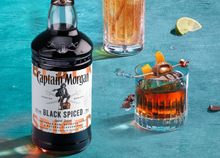 Bottle of Captain Morgan Black Spiced Rum on blue background beside a short glass of dark liquid and a tall glass of gold liquid.