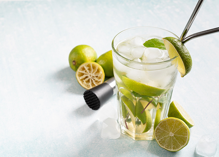 One glass of a vodka cocktail with mint and lime wedges, staged next to cocktail equipment and more limes on a white surface.