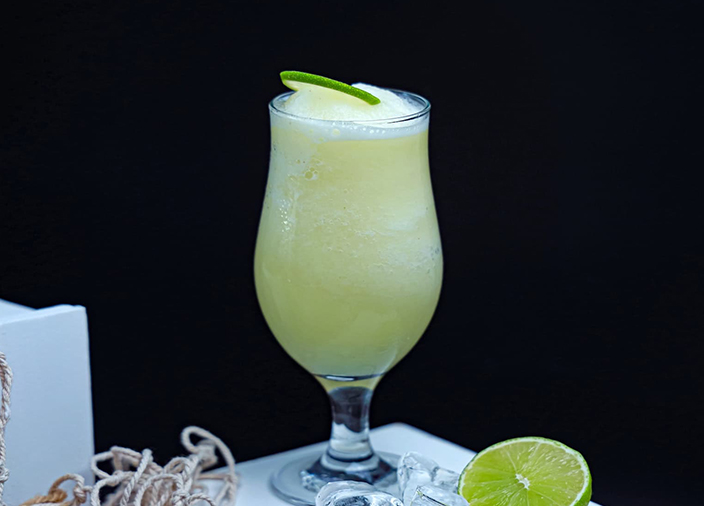 A Gin Rickey in a tall highball glass filled with ice and lime wedge staged on a wooden table. Surrounded by ingredients: ice cubes, lime wedges, ice and herbs.