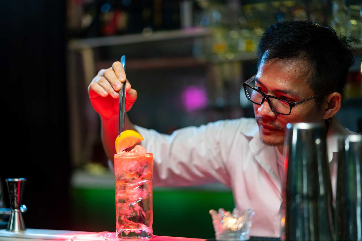 Um bartender de óculos e camisa branca usa pinças para colocar uma rodela de limão em um copo alto cheio de gelo e drink âmbar efervescente. A cena se passa em um bar com luzes vermelhas e rosa desfocadas ao fundo.