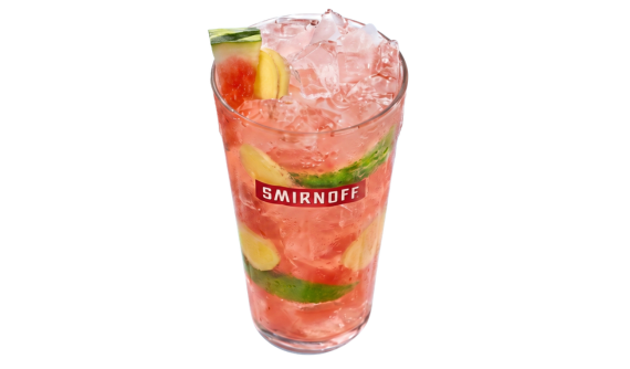 Copo alto com logo vermelho da Smirnoff contendo um coquetel rosa com bastante gelo. No interior da bebida, há fatias de pepino e gengibre. A borda é decorada com um pedaço triangular de melancia e uma rodela de gengibre. Fundo branco.