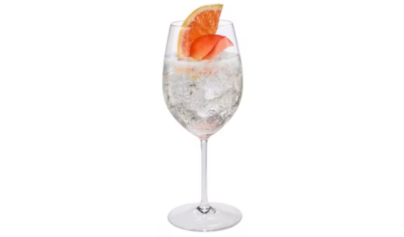 Taça de vinho de haste longa contendo um coquetel claro e borbulhante cheio de gelo triturado. A bebida refrescante é decorada no topo com uma fatia de toranja rosada e uma pétala de flor avermelhada. Fundo branco isolado.