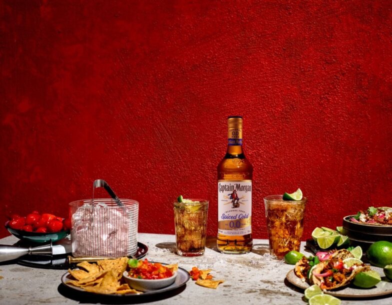 Bottle of Captain Morgan Spiced Gold 0.0% Alcohol Free Spirit on table surrounded by assorted foods and drinks against a red background