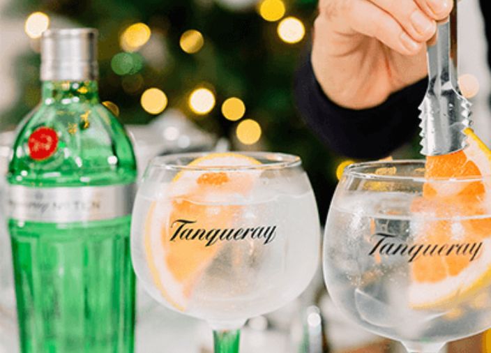 Dois cocktails de Gin com um bartender acrescentando uma rodela de laranja a uma das bebidas. Uma garrafa de Tanqueray No.10 está ao fundo