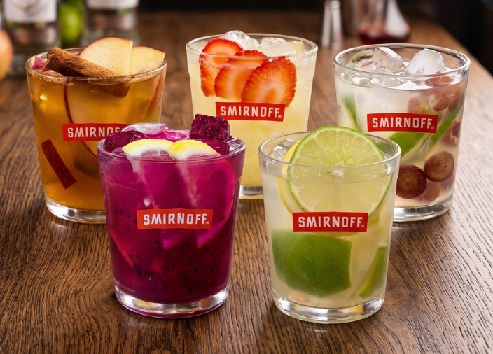 Cinco copos baixos com logo da Smirnoff sobre um balcão de madeira, contendo coquetéis coloridos com gelo e frutas frescas: pitaya, limão, maçã com canela, morango e uva. Ao fundo desfocado, garrafas de vodka e um ambiente de bar.