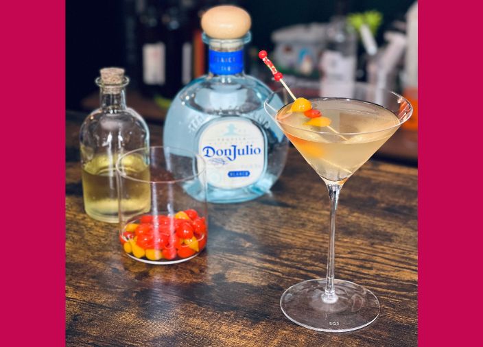 Ein orangefarbener Martini-Cocktail mit einer roten und orangefarbenen Gurkengarnitur und einer Flasche Don Julio Tequila auf einem Holztisch.