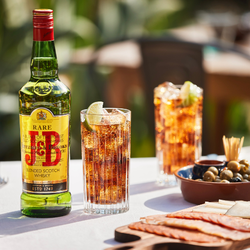 Botella de whisky J&B en una mesa exterior con dos cócteles y un plato de aceitunas.