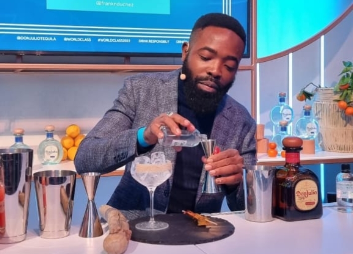 A man standing behind a bar with a head mounted microphone pouring a small bottle of clear liquid into a measuring shot next to a glass with ice.