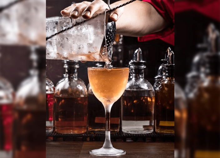 Bartenders hand pouring an orange-coloured cocktail into a glass from a strainer.