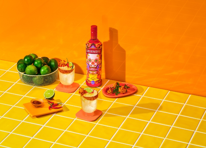 A bottle of Smirnoff Spicy Tamarind beside a cocktail, plate of spicy food and bowl of limes against an orange background.
