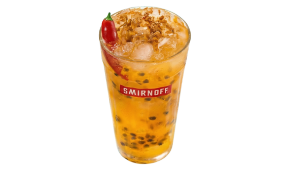 Copo alto com logo vermelho da Smirnoff contendo um coquetel amarelo de maracujá com sementes e gelo. Uma pimenta vermelha inteira decora a lateral e há especiarias polvilhadas no topo. Fundo branco isolado.
