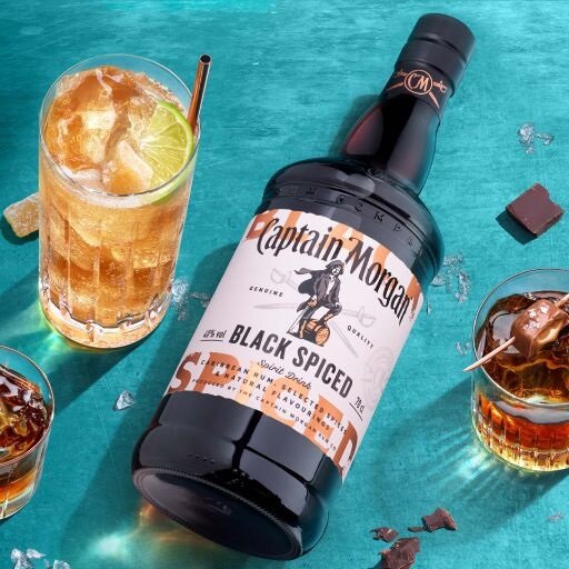 Bottle of Captain Morgan Black Spiced Rum on blue background beside a short glass of dark liquid and a tall glass of gold liquid.