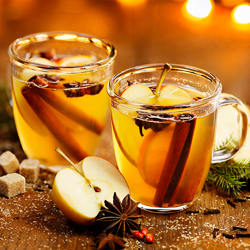 Two Toffee Apple Toddy cocktails in glass cups garnished with apple slices, a cinnamon stick and whole star anise. All surrounded by more garnish and a sprig of holly, sugar cubes and fairy lights. 