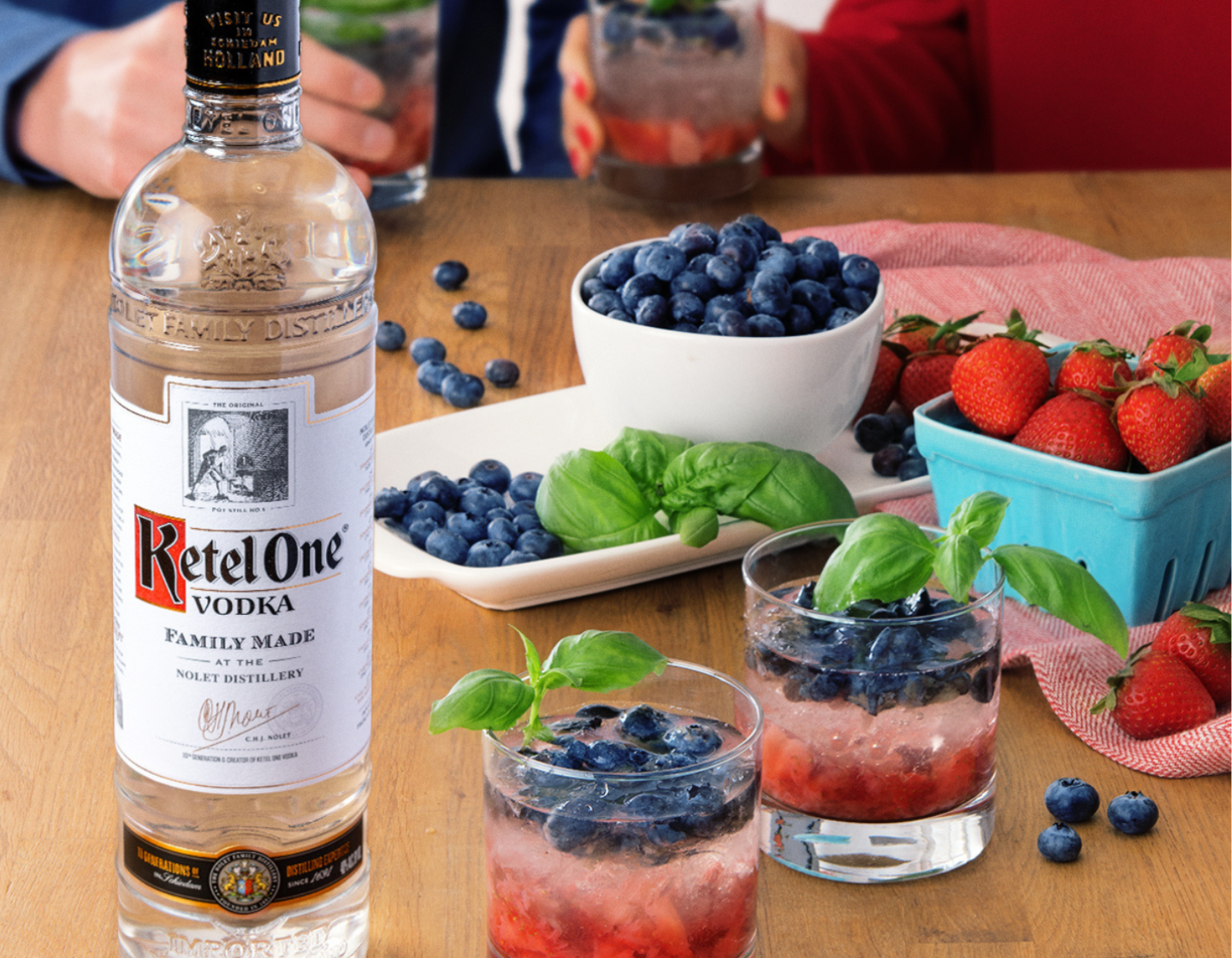 Garrafa de vodka Ketel One ao lado de dois coquetéis recheados de mirtilos, ao lado de tigelas com frutas variadas.