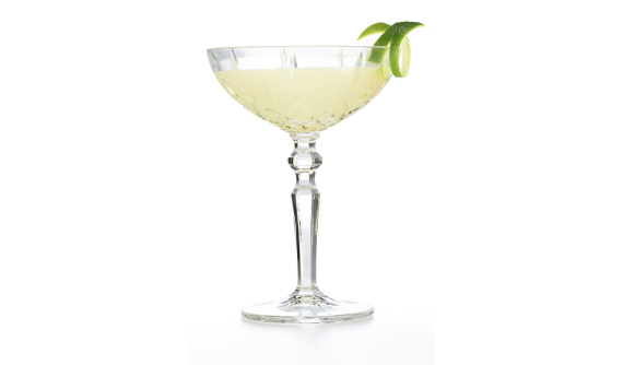 Coquetel de cor amarelo-pálido servido em uma taça coupe de cristal lapidado com haste elegante. Uma casca de limão verde em espiral decora a borda direita. O fundo é totalmente branco, isolando a bebida.