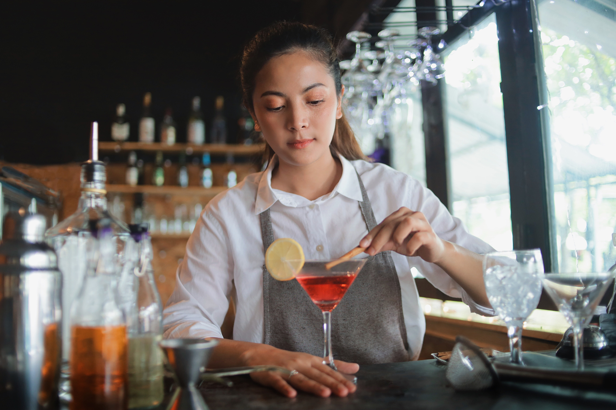 A female bartender in an apron decorates a red cocktail in a martini glass with a cinnamon stick. The glass already has a slice of lemon on the rim. Bar utensils such as a cocktail shaker and jigger are on the counter.