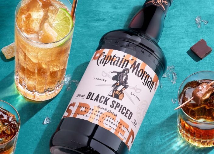 Botella de ron Captain Morgan Black Spiced sobre fondo azul junto a un vaso corto de líquido oscuro y un vaso alto de líquido dorado.