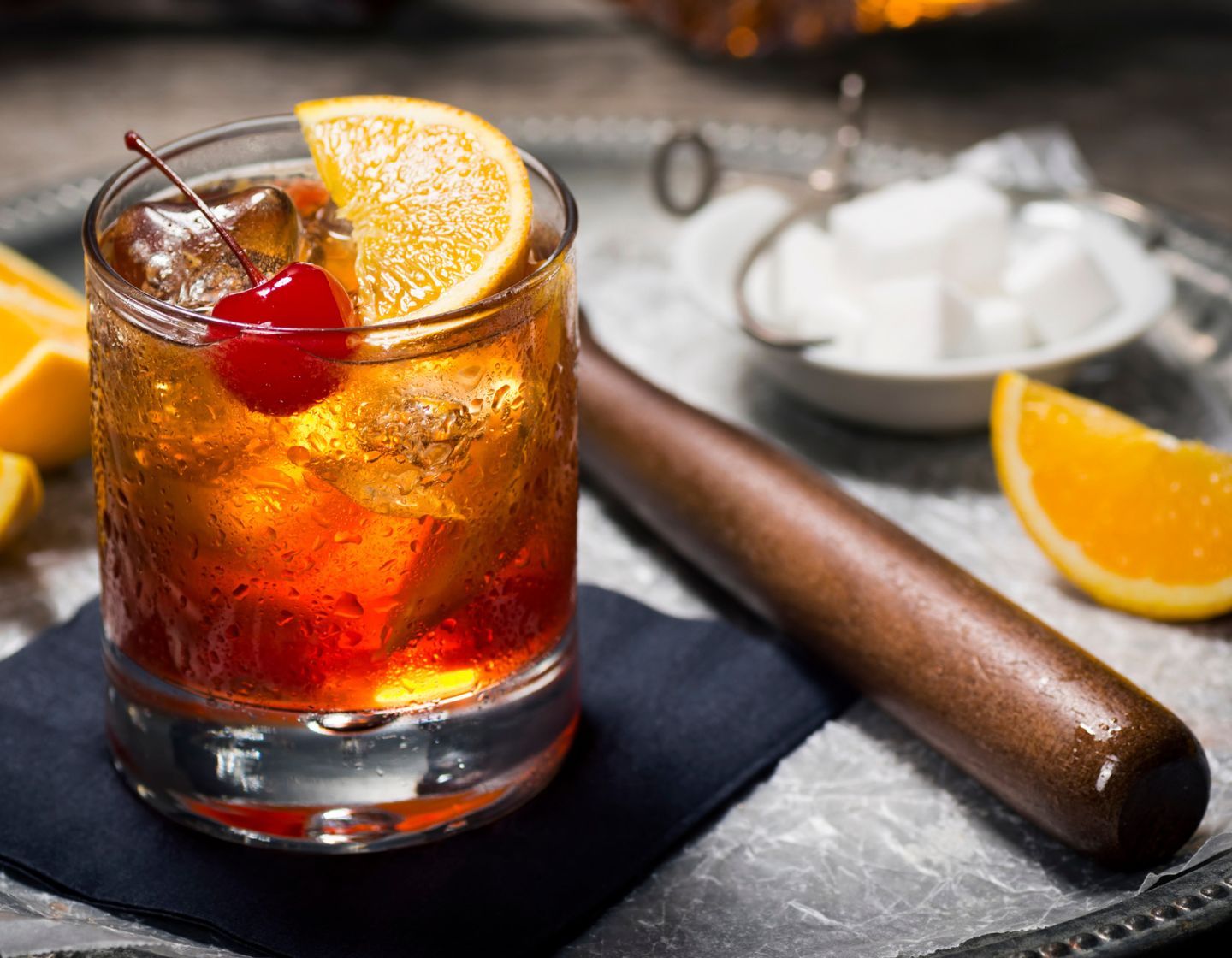 Herramientas de maceración en una encimera, con un cóctel de naranja adornado con naranja y cereza.