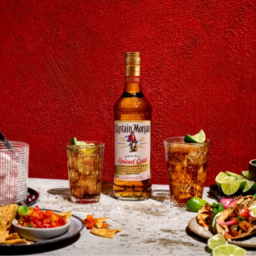 Bottle of Captain Morgan Spiced Gold rum on table beside two iced glasses of rum and coke surrounded by food