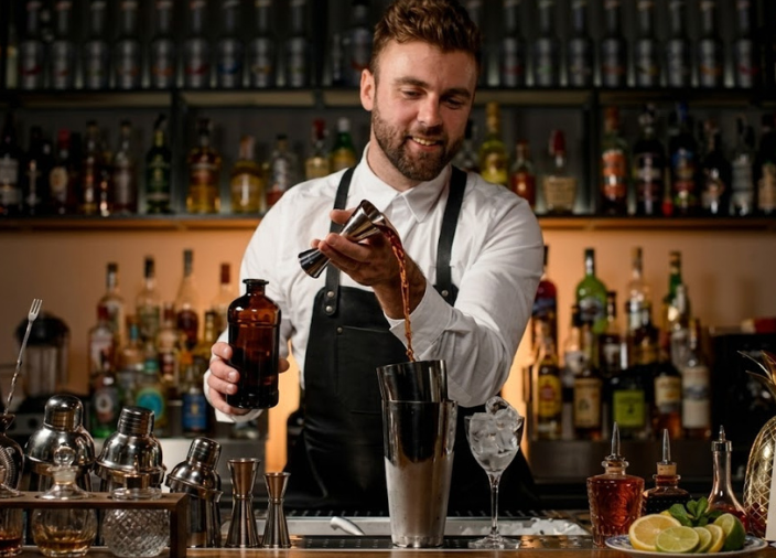 Bartender sorridente de camisa branca e avental preto prepara um coquetel, despejando bebida de um jigger para uma coqueteleira de metal. O balcão exibe utensílios de bar e um abacaxi dourado. Ao fundo, prateleiras iluminadas com garrafas.