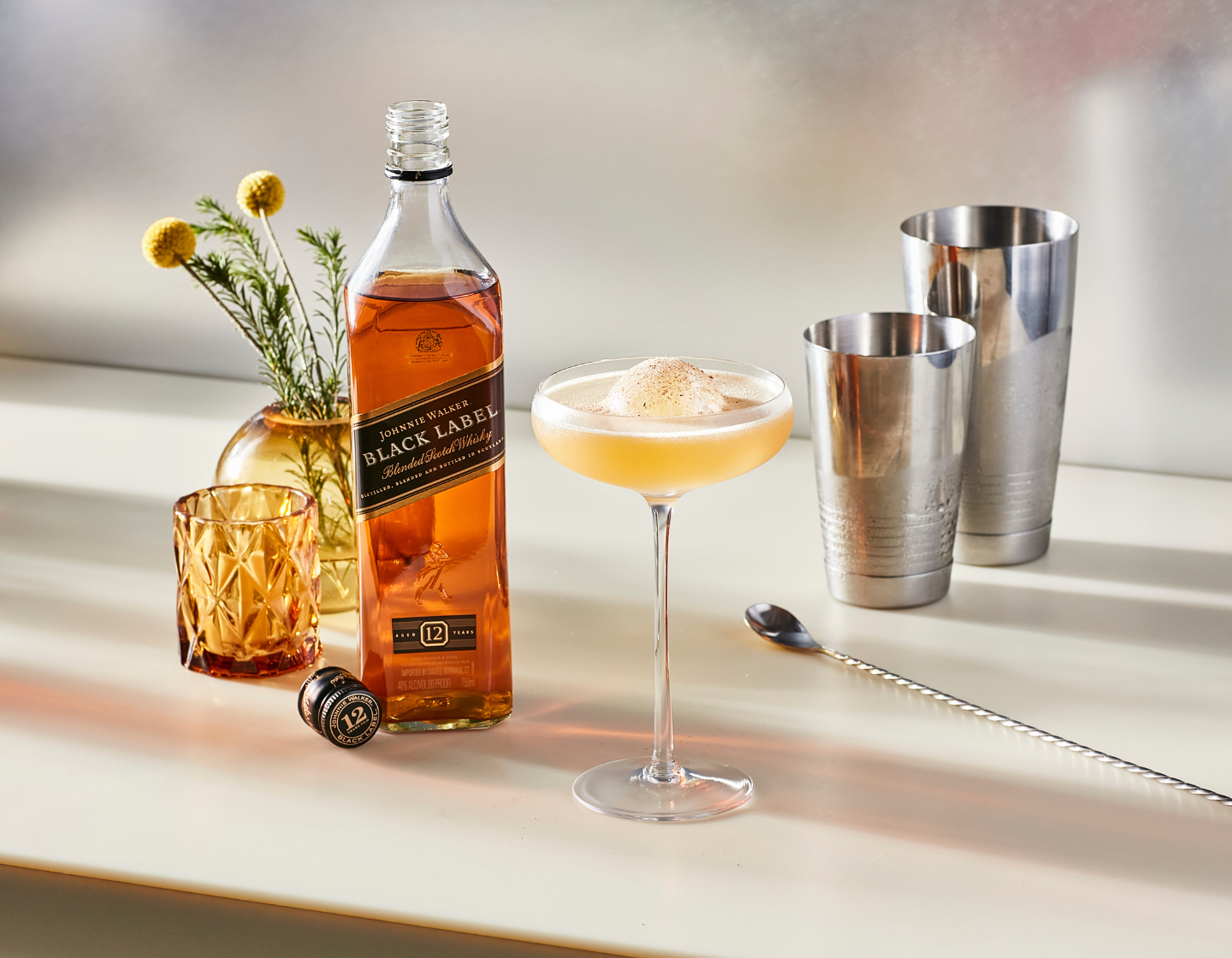 Cocktail in a coupe glass with ice, surrounded by bottle of Johnnie Walker Black Label Whisky, cocktail shaker and bar spoon.