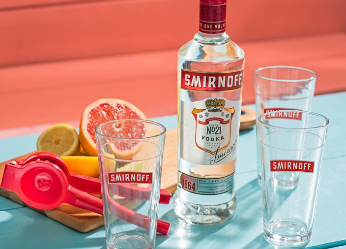Garrafa de vodka Smirnoff No. 21 e três copos vazios da marca sobre uma mesa de madeira azul-clara sob luz solar. Ao lado, uma tábua de corte exibe toranja e limão fatiados junto a um espremedor vermelho. Fundo coral vibrante.