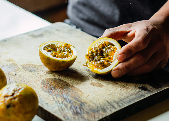 One hand holding one half of a sliced open passion fruit on a chopping board, the inside seeds showing.