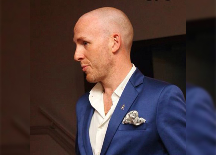 Perfil lateral de Tim Etherington-Judge vestindo um terno azul-marinho e camisa branca. 