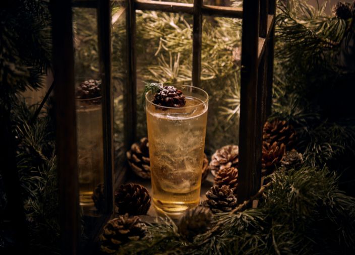 A highball cocktail surrounded by pinecones topped with a chocolate pinecone garnish