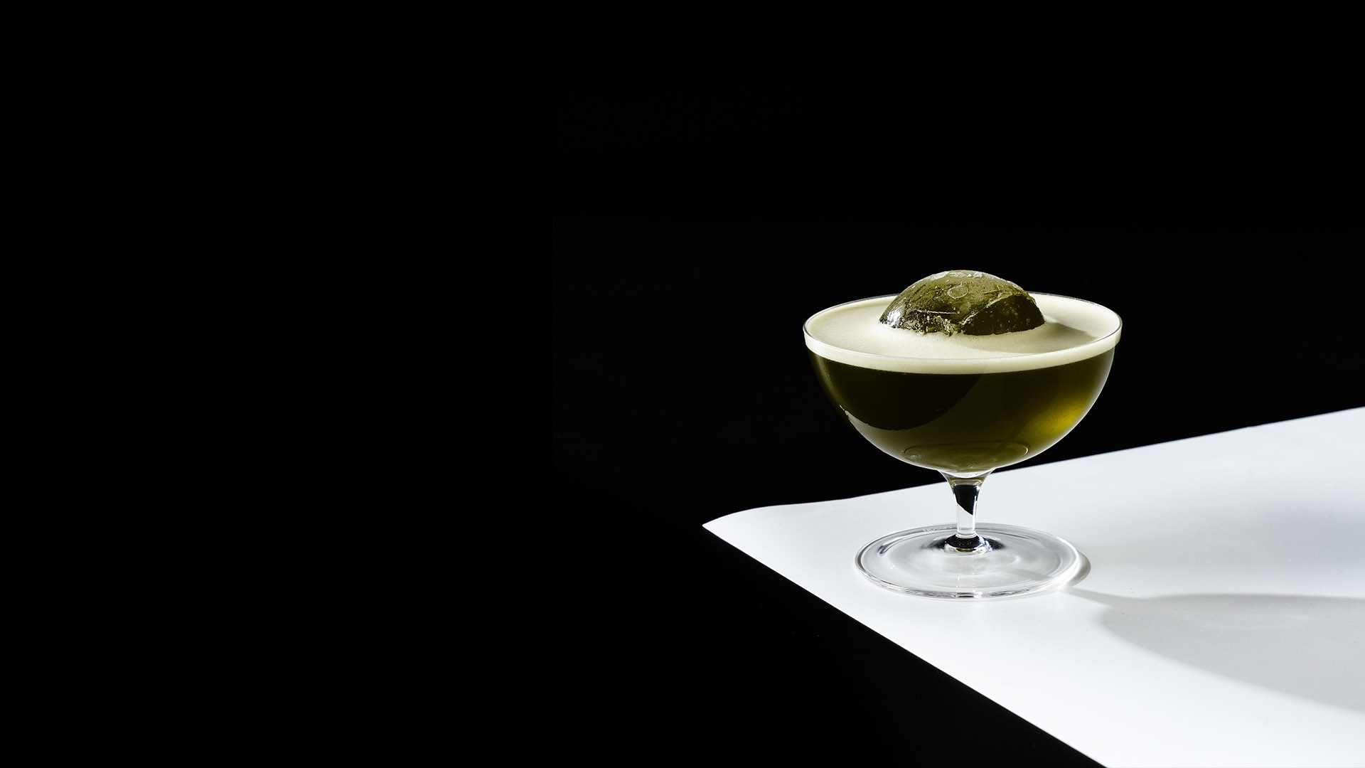 Coquetel verde-escuro servido em taça coupe. Possui uma camada de espuma clara e uma grande esfera de gelo translúcida em destaque no centro. A taça repousa sobre uma superfície branca diagonal que contrasta fortemente com o fundo totalmente preto.