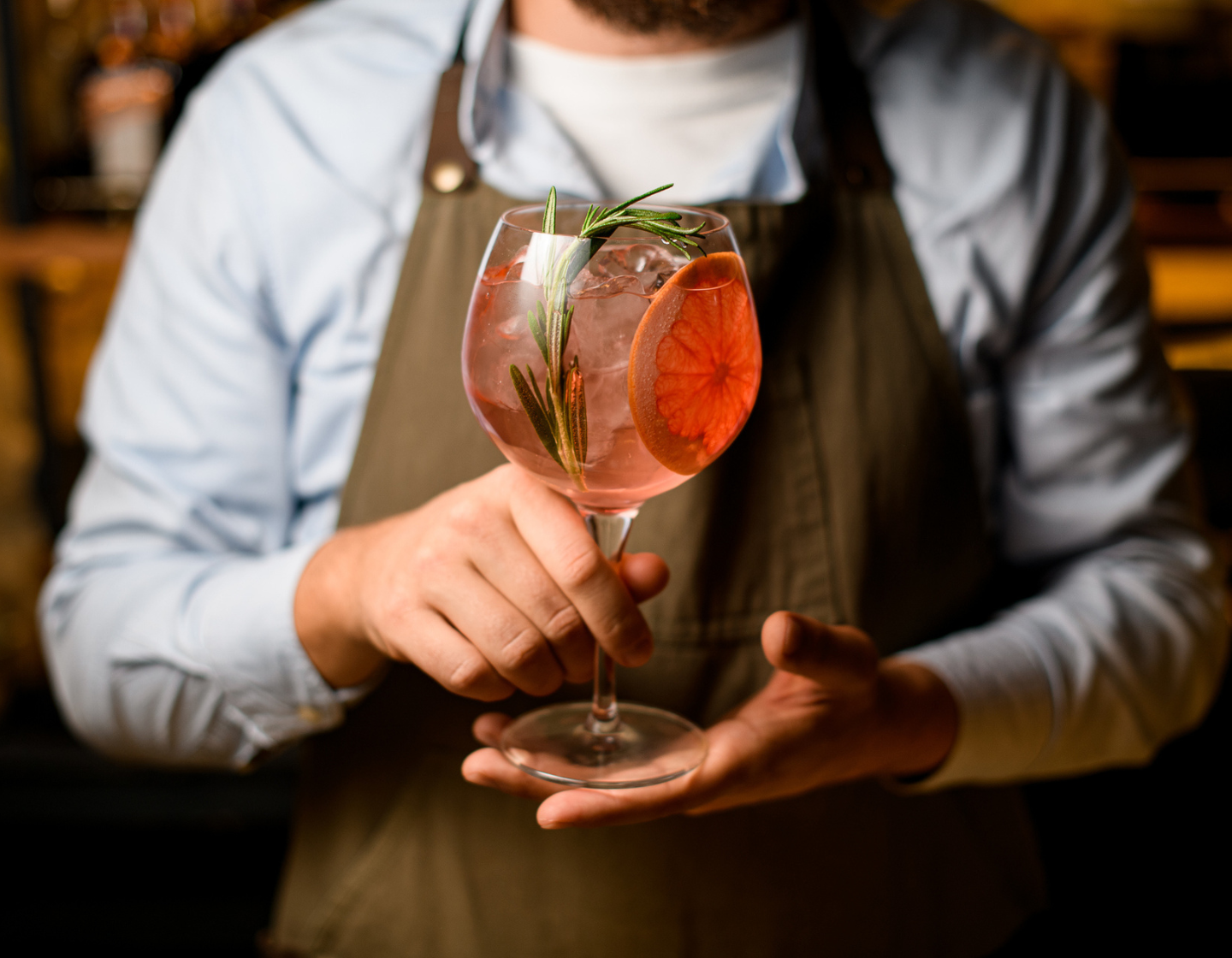 Hombre con delantal sosteniendo un servicio de ginebra con guarnición de naranja en un vaso grande con hielo y romero. 