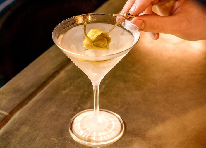 A martini drink with a bartender’s hand adding a green pickle garnish.