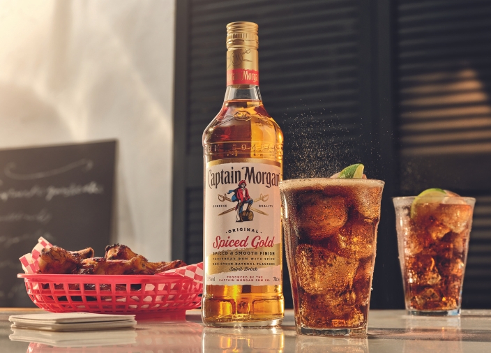 Botella de Captain Morgan Original Spiced Gold sobre una mesa, rodeada de diferentes platos y bebidas, sobre un fondo rojo
