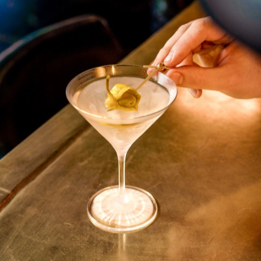 A martini drink with a bartender’s hand adding a green pickle garnish.