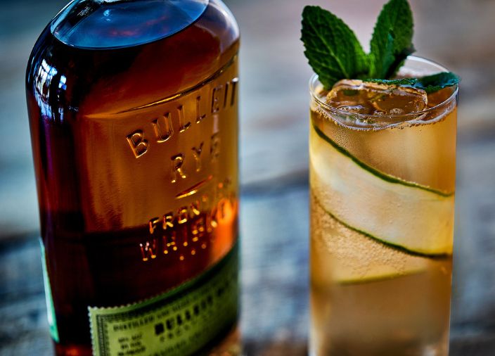 Coquetel de whiskey Bulleit bourbon em copo highball decorado com fatia de pepino e garrafa de bourbon Bulleit 