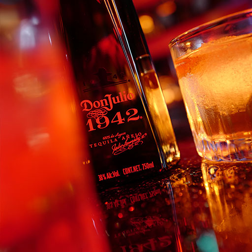 Bottle of Don Julio 1942 tequila against gold curtain beside glass of liquid and ice