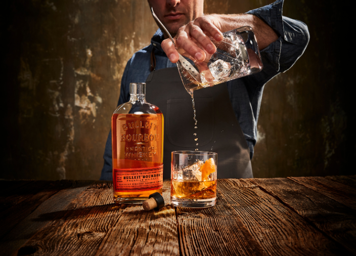 Mann, der einen Cocktail in ein Glas auf dem Tisch neben einer offenen Flasche Bulleit Whiskey abseiht