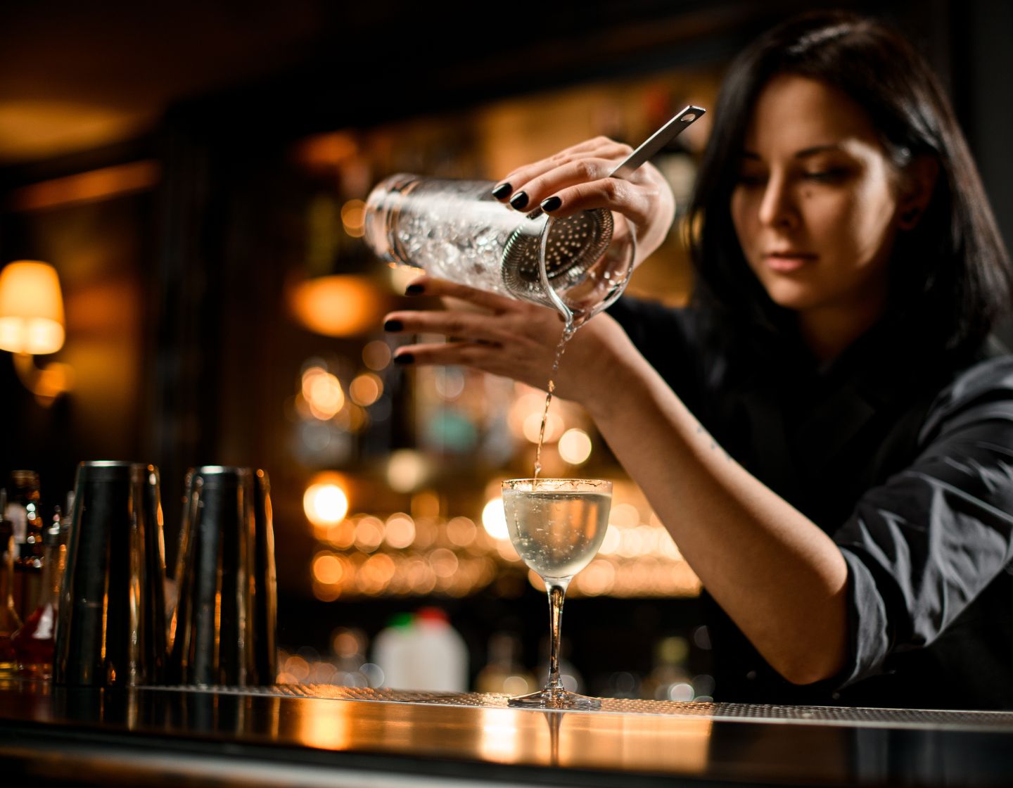 Female bartender pouring cocktail through a strainer into a classic coupe cocktail glass.