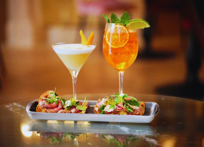 A yellow cocktail and an orange cocktail with a plate of food.