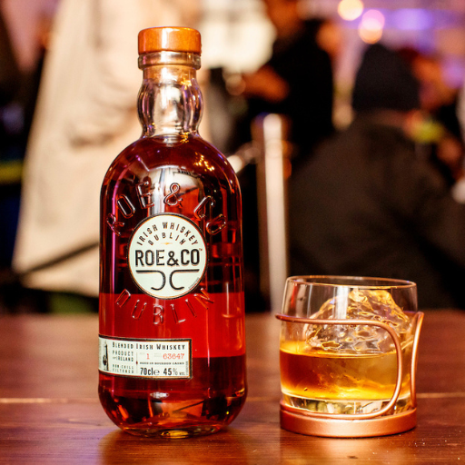 Botella de whisky Roe & Co en una mesa junto a un vaso de whisky con hielo.