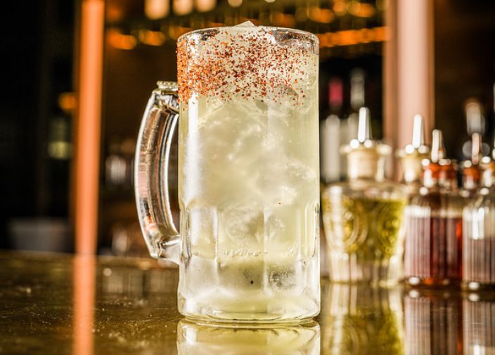 A yellow-tinted cocktail in a stein glass with a red, salted rim.