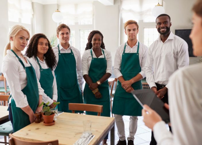 Six bartenders in aprons being addressed by a manager