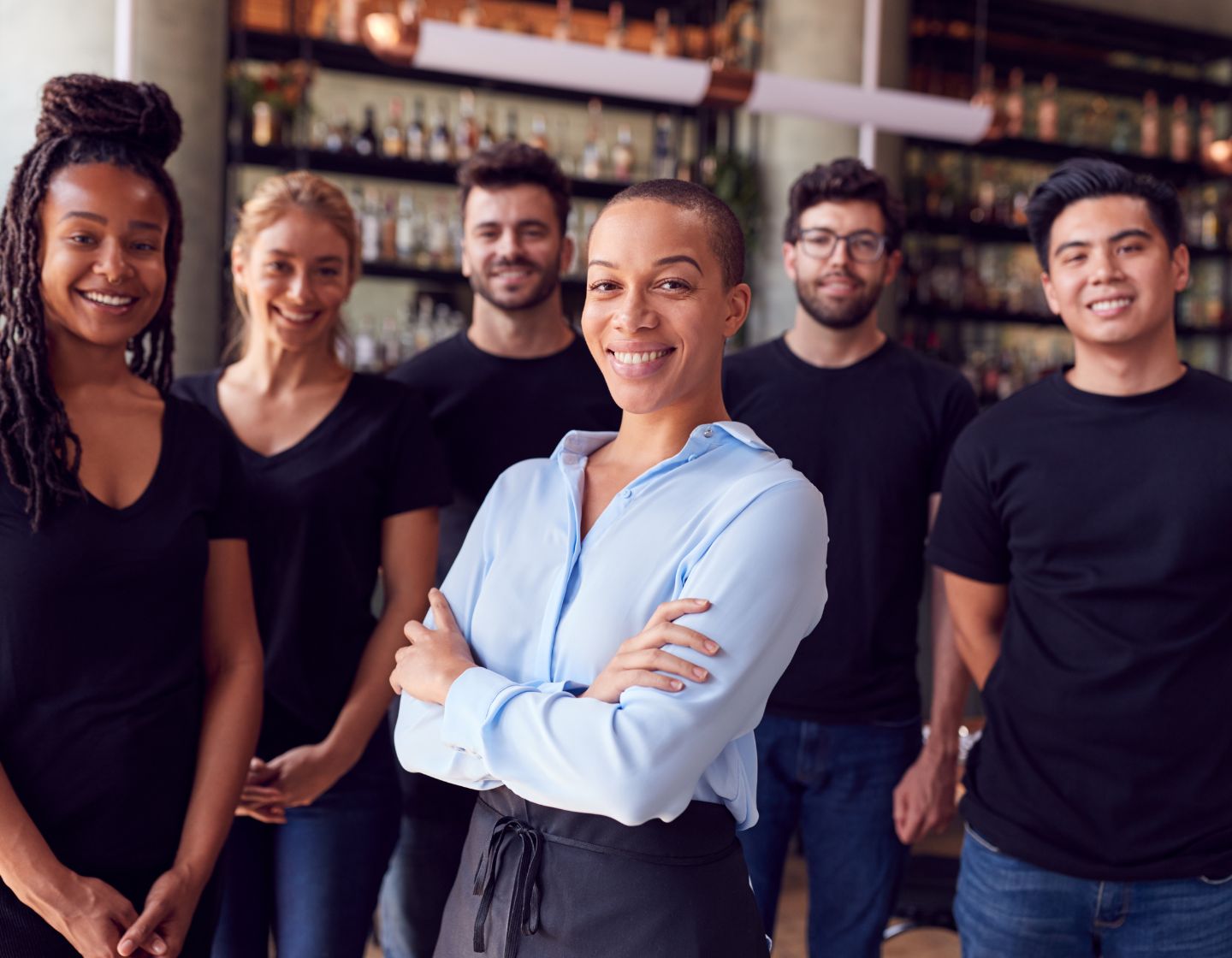 Una gerente de bar de pie con su equipo, con los brazos cruzados. El equipo está un poco atrás, sonriendo.