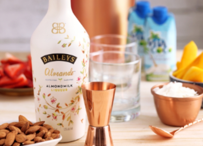 Una botella de Baileys Almande, un licor vegano, sobre una mesa con un bol de almendras, un bol de melocotón en rodajas, un bol de fresas en rodajas, dos vasos y utensilios para preparar cócteles.