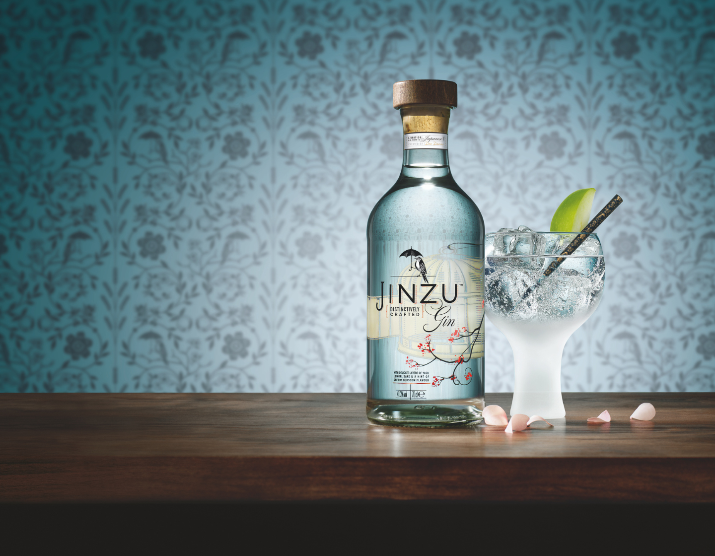 Bottle of Jinzu gin on table beside glass of gin and tonic against blue floral wallpaper in background