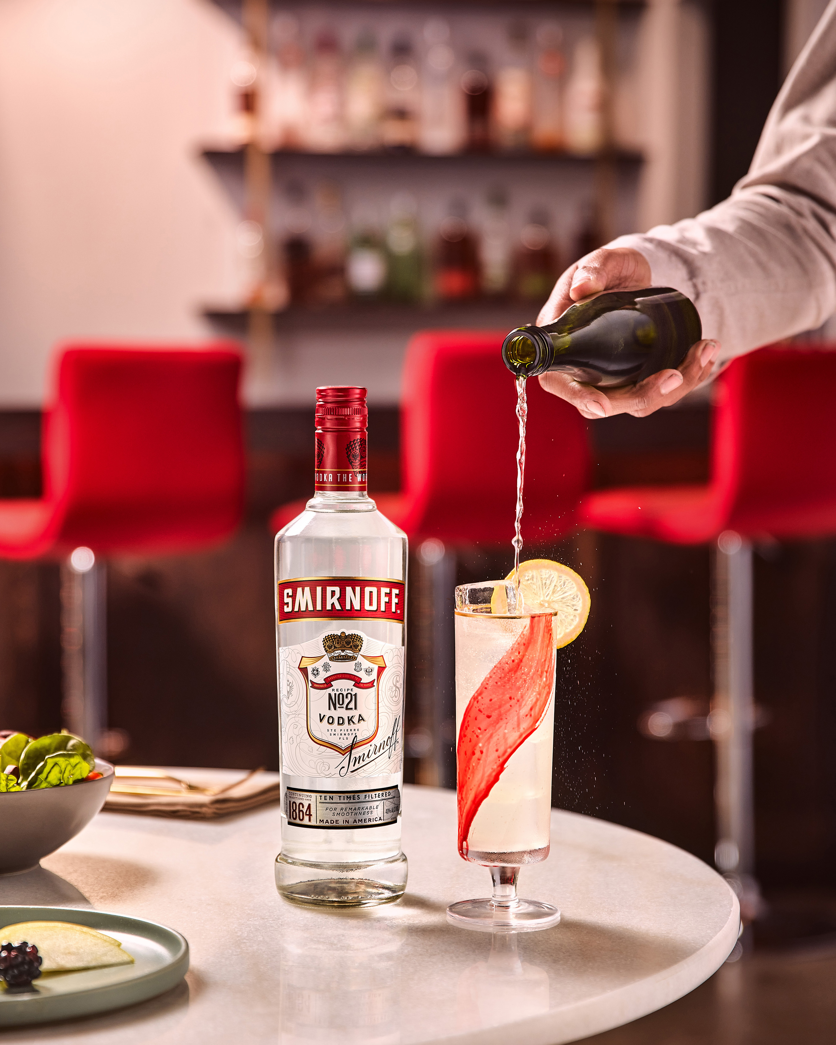 Garrafa de Smirnoff No. 21 sobre mesa de mármore. Ao lado, uma mão serve líquido de uma garrafa verde pequena em uma taça alta com gelo, decorada com uma fita vermelha interna e rodela de limão. Fundo de bar desfocado com cadeiras vermelhas.