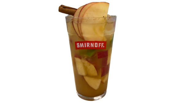 Copo alto com logo vermelho da Smirnoff contendo um coquetel dourado com pedaços de maçã mergulhados. O topo da bebida é elegantemente decorado com fatias finas de maçã em leque e um pau de canela rústico. Fundo branco isolado.