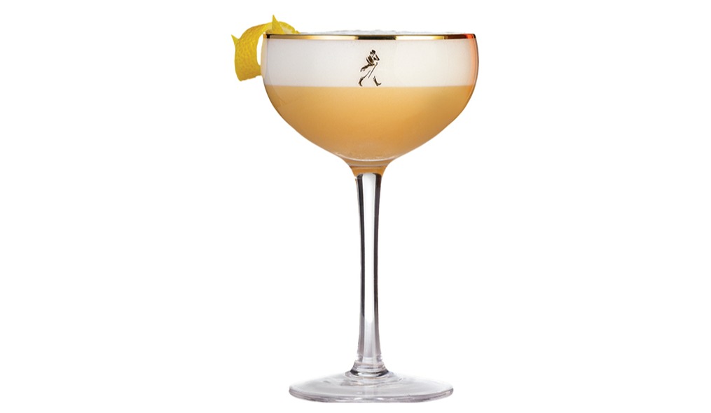 Taça coupe de haste longa com borda dourada e logo preto do Johnnie Walker. Contém um coquetel com base amarelo-âmbar e uma espessa camada de espuma branca no topo. Uma casca de limão fina decora a borda esquerda. Fundo branco.
